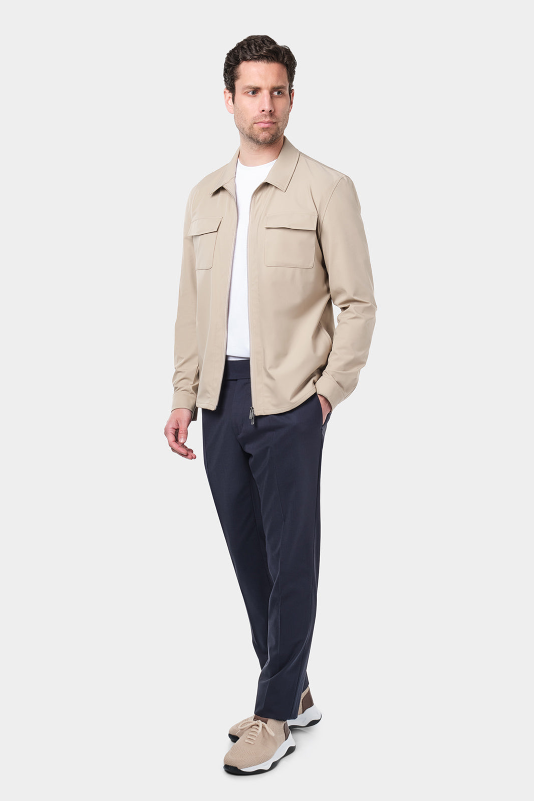 Un homme vêtu d'un blouson beige en tissu Olmetex, imperméable et coupe-vent, décontracté, avec quatre poches, une fermeture à double zip et un col. Il porte un tee-shirt blanc en coton glacé et un pantalon bleu marine à pinces, accompagné de sneakers beiges en maille et cuir.