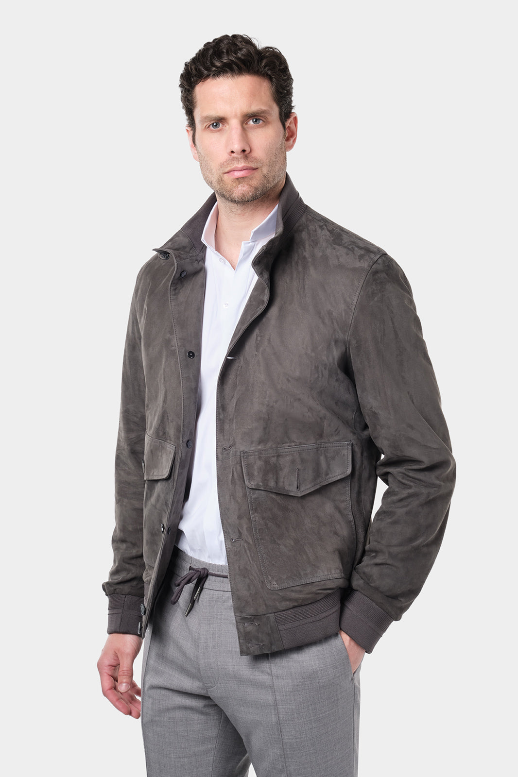 Un homme aux cheveux courts et foncés porte une chemise blanche à col inversé en tissu confortable de coton et élasthanne, assortie à un pantalon jogpant gris clair avec un cordon à la taille. Il porte un blouson en daim gris foncé avec des fermetures à boutons et de grandes poches ainsi que des baskets beiges en maille et en cuir et se tient debout contre un fond gris clair uni, regardant droit devant lui.