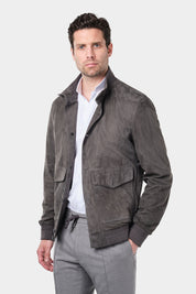 Un homme aux cheveux courts et foncés porte une chemise blanche à col inversé en tissu confortable de coton et élasthanne, assortie à un pantalon jogpant gris clair avec un cordon à la taille. Il porte un blouson en daim gris foncé avec des fermetures à boutons et de grandes poches ainsi que des baskets beiges en maille et en cuir et se tient debout contre un fond gris clair uni, regardant droit devant lui.