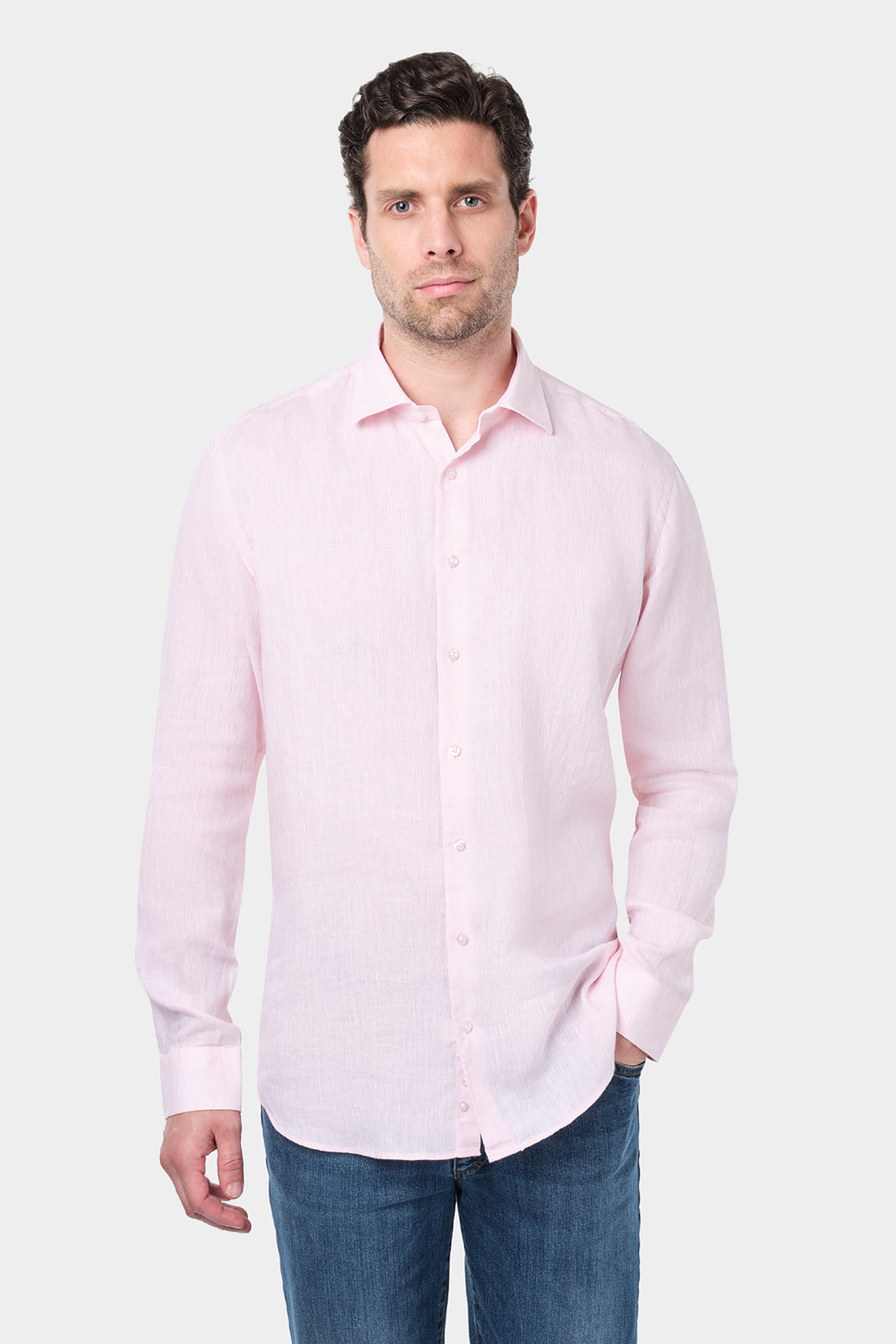 Un homme aux cheveux courts et foncés porte une chemise rose à col traditionnel en lin. Il est assorti à un jean bleu. Il se tient debout contre un fond gris clair uni, regardant droit devant lui.