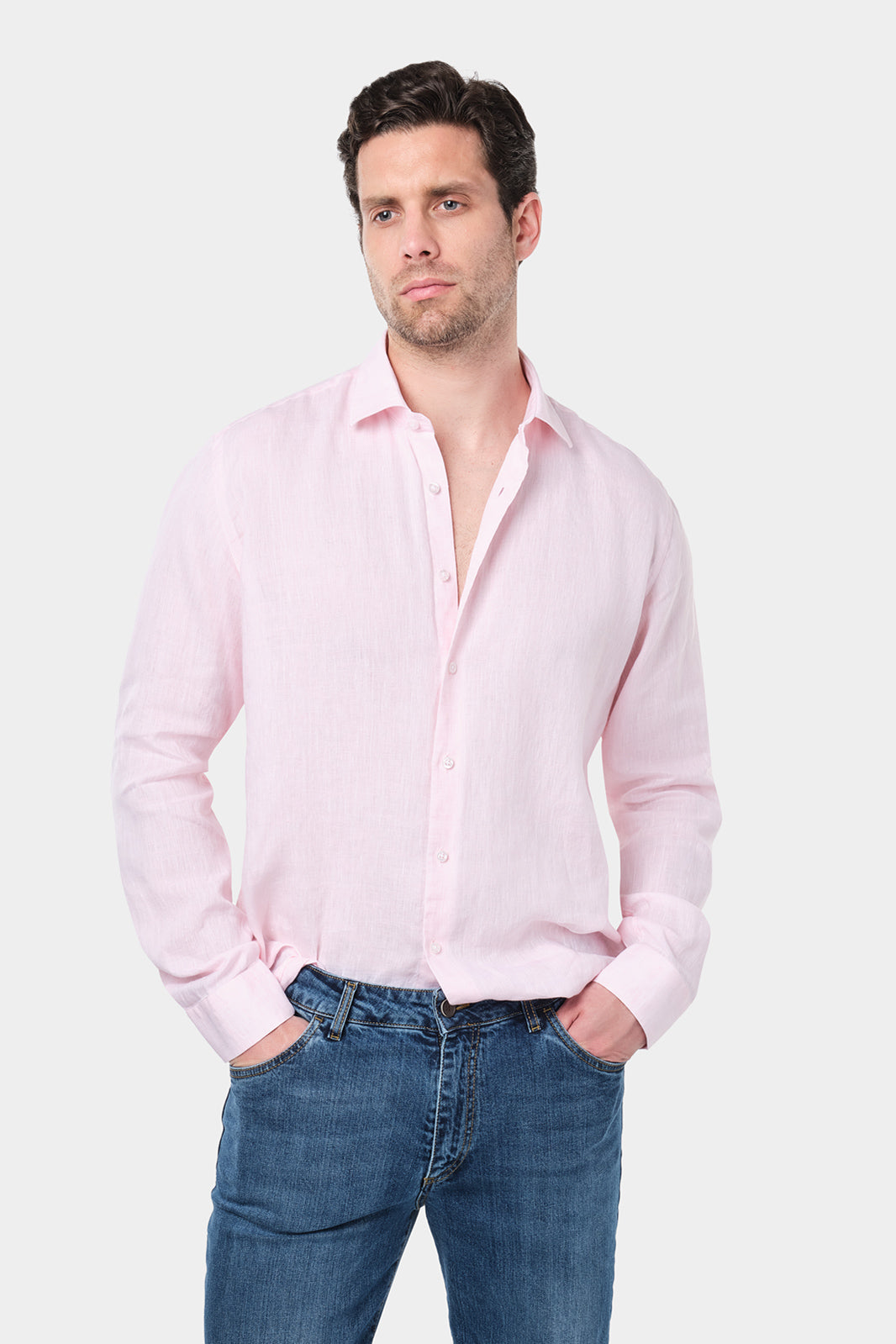 Un homme aux cheveux courts et foncés porte une chemise rose à col traditionnel en lin. Il est assorti à un jean bleu. Il se tient debout contre un fond gris clair uni.