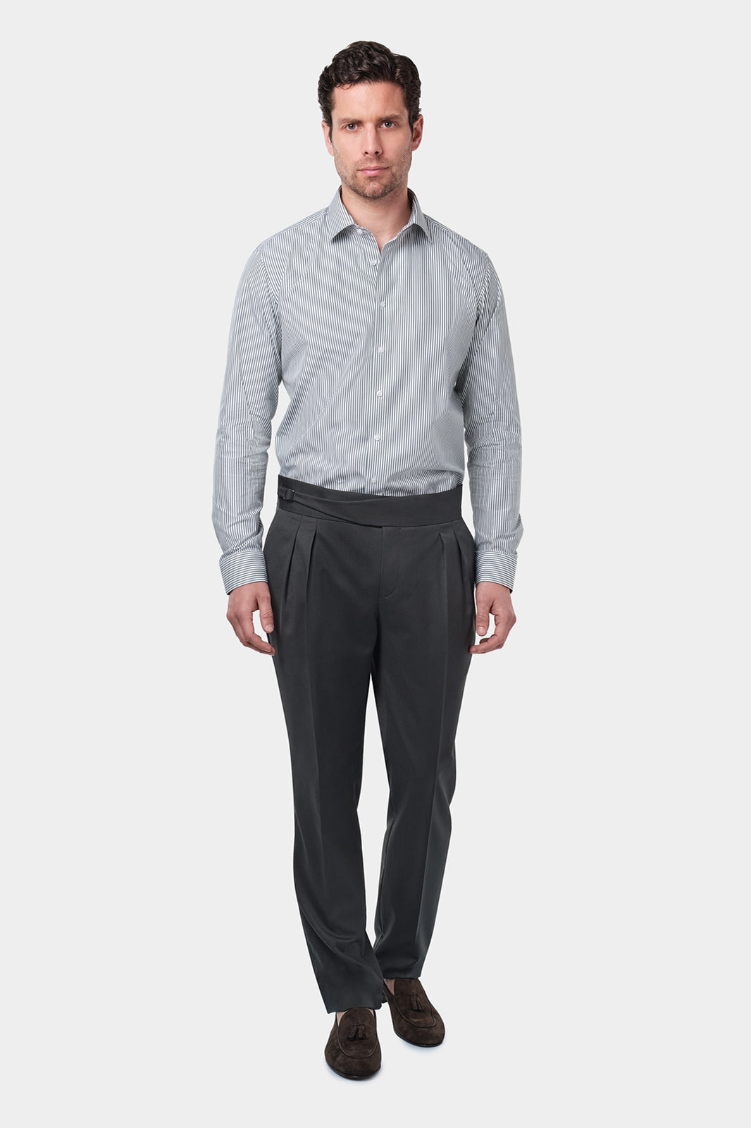 Un homme vêtu d'une chemise à rayures kaki et blanches avec un col français. Il porte également un pantalon gris foncé à pinces, accompagné de mocassins en daim marron.L'homme se tient debout, les mains détendues le long du corps, sur un fond gris clair uni.