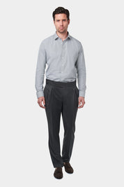 Un homme vêtu d'une chemise à rayures kaki et blanches avec un col français. Il porte également un pantalon gris foncé à pinces, accompagné de mocassins en daim marron.L'homme se tient debout, les mains détendues le long du corps, sur un fond gris clair uni.