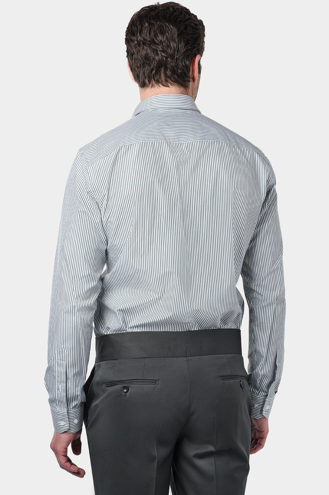 Un homme de dos vêtu d'une chemise à rayures kaki et blanches avec un col français. Il porte également un pantalon gris foncé à pinces, accompagné de mocassins en daim marron.L'homme se tient debout, les mains détendues le long du corps, sur un fond gris clair uni.