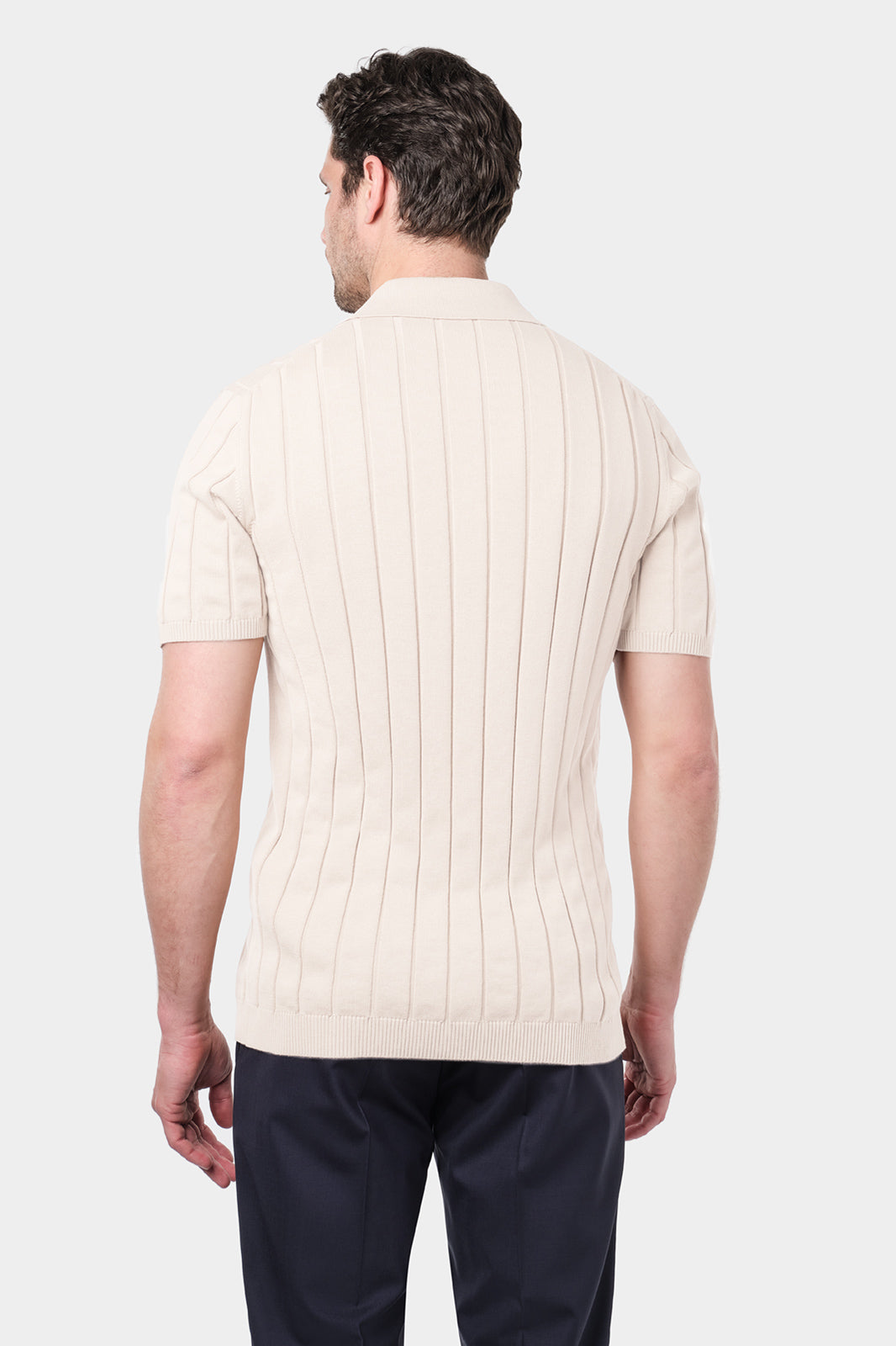 Un homme de dos portant une chemise polo beige en coton avec des rayures verticales et un pantalon bleu marine.