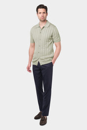 Un homme portant une chemise polo sauge en coton avec des rayures verticales et un pantalon bleu marine. Il a les mains dans les poches et porte des mocassins en daim marron. Le fond est uni et de couleur claire.