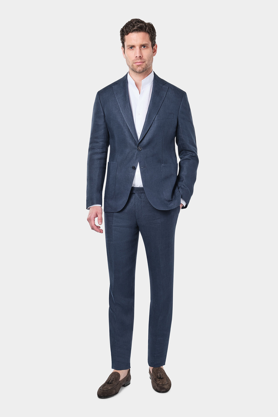 Un homme porte un costume en lin de couleur marine, accompagné d'une chemise blanche et de mocassins marron. Il se tient debout avec une main dans la poche, dans un style élégant et décontracté.
