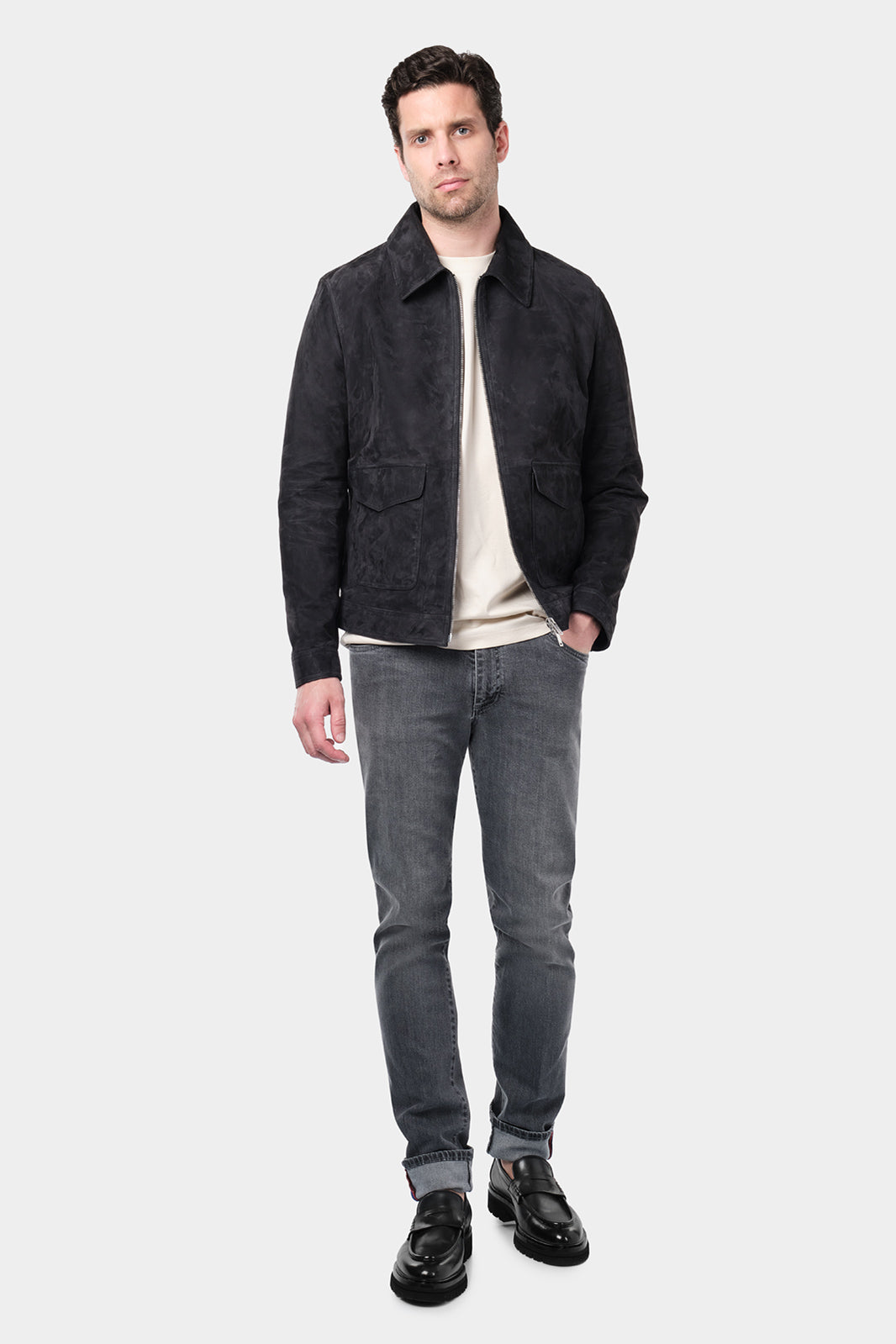 Un homme portant un jean coupe slim en coton et élasthanne de couleur grise. Il porte également un tee-shirt en coton glacé beige, un blouson en peau et des mocassins en cuir noir.