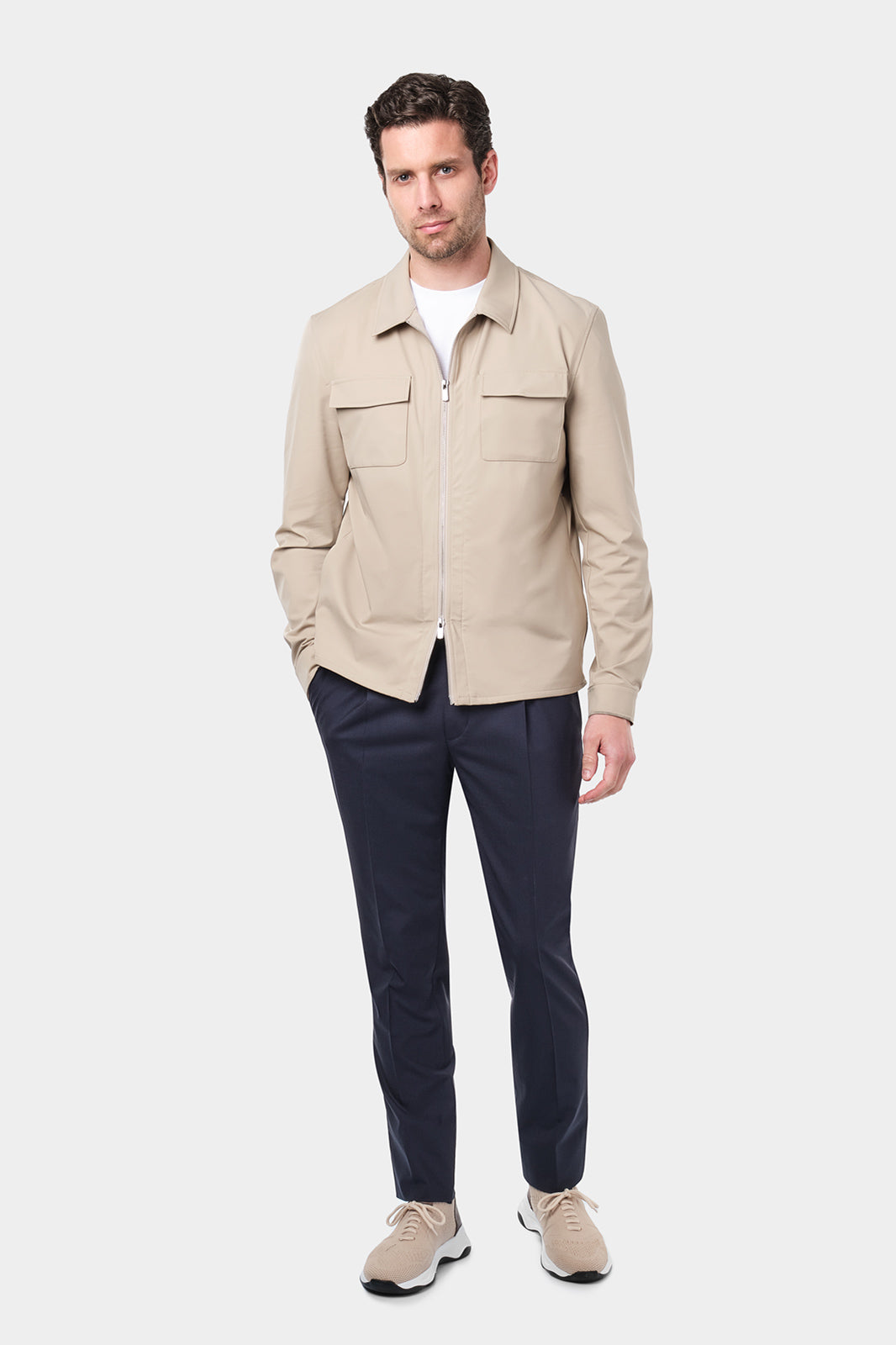 Un homme vêtu d'un blouson beige en tissu Olmetex, imperméable et coupe-vent, décontracté, avec quatre poches, une fermeture à double zip et un col. Il porte un tee-shirt blanc en coton glacé et un pantalon bleu marine à pinces, accompagné de sneakers beiges en maille et cuir.