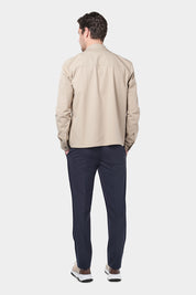 Un homme de dos vêtu d'un blouson beige en tissu Olmetex, imperméable et coupe-vent, décontracté, avec quatre poches, une fermeture à double zip et un col. Il porte un tee-shirt blanc en coton glacé et un pantalon bleu marine à pinces, accompagné de sneakers beiges en maille et cuir.