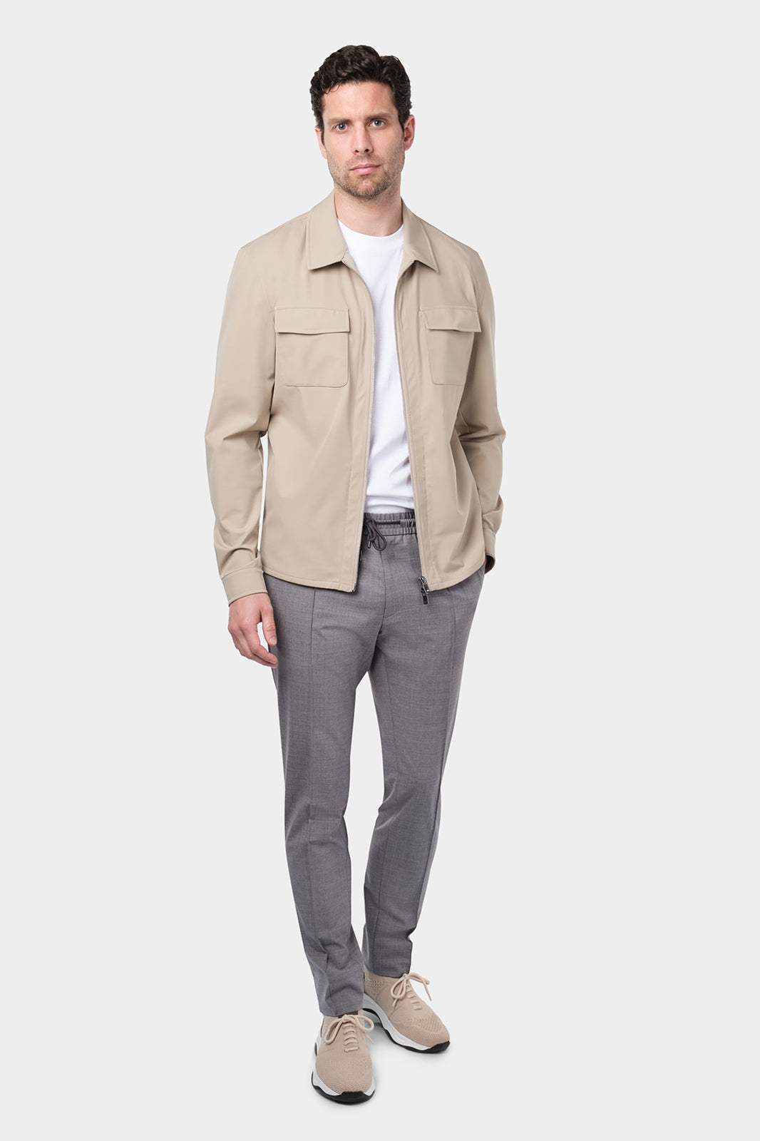 Un homme vêtu d'un blouson beige en tissu Olmetex, imperméable et coupe-vent, décontracté, avec quatre poches, une fermeture à double zip et un col. Il porte un tee-shirt blanc en coton glacé et un pantalon bleu marine à pinces, accompagné de sneakers beiges en maille et cuir.