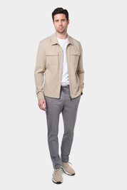 Un homme vêtu d'un blouson beige en tissu Olmetex, imperméable et coupe-vent, décontracté, avec quatre poches, une fermeture à double zip et un col. Il porte un tee-shirt blanc en coton glacé et un pantalon bleu marine à pinces, accompagné de sneakers beiges en maille et cuir.