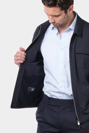  Un homme vêtu d'un blouson bleu marine en tissu Olmetex, imperméable et coupe-vent, décontracté, avec quatre poches, une fermeture à double zip et un col. Il porte une chemise à rayures bleu ciel et blanches avec un col français et un pantalon bleu marine à pinces.