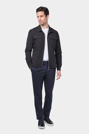 Un homme vêtu d'un blouson noir en tissu Olmetex, imperméable et coupe-vent, décontracté, avec quatre poches, une fermeture à double zip et un col. Il porte un tee-shirt blanc en coton glacé et un pantalon bleu marine à pinces, accompagné de sneakers en bi matière veau velours.