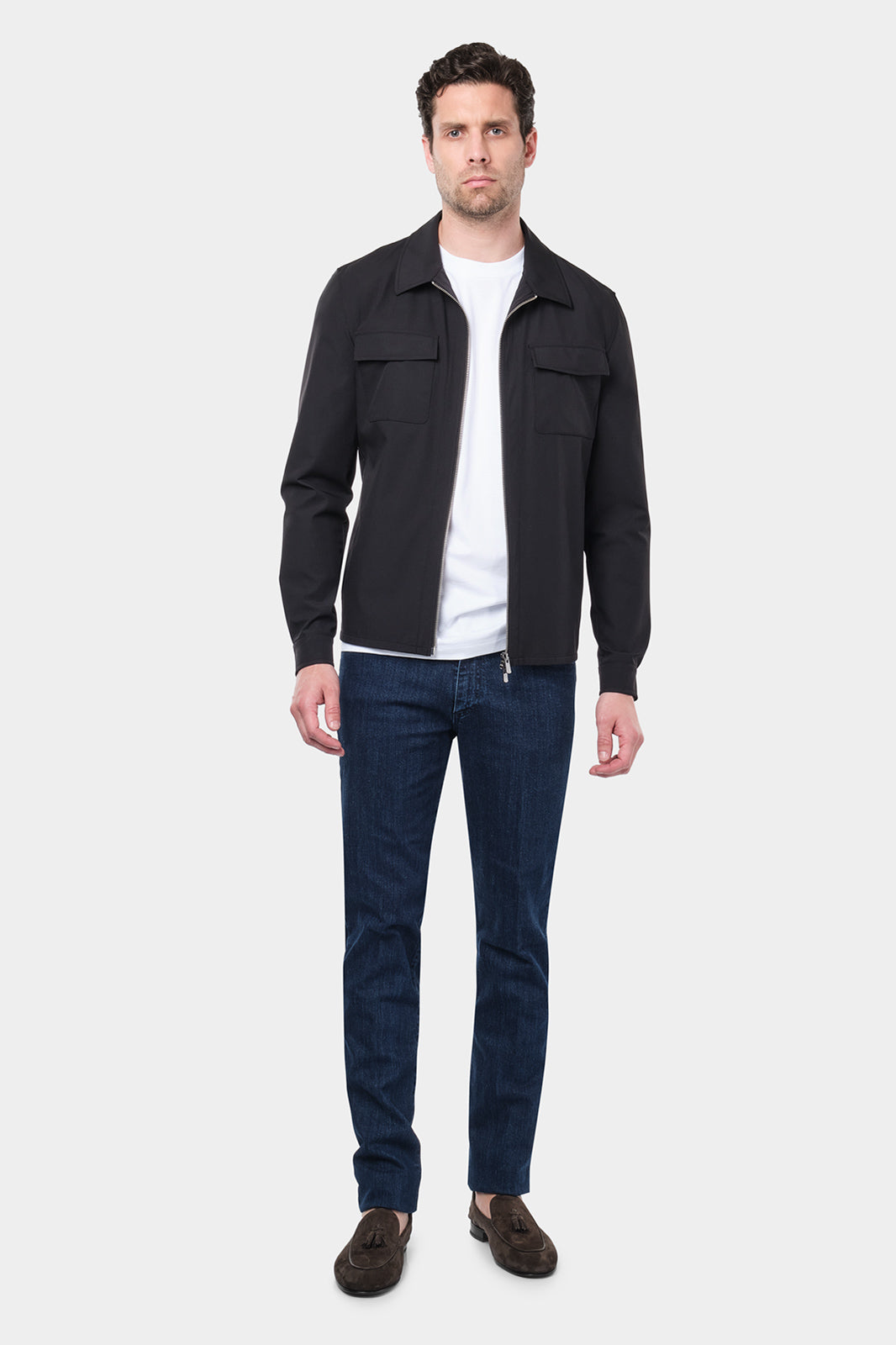 Un homme vêtu d'un blouson noir en tissu Olmetex, imperméable et coupe-vent, décontracté, avec quatre poches, une fermeture à double zip et un col. Il porte un tee-shirt blanc en coton glacé et un jean couleur brut accompagné de mocassins en daim marron.