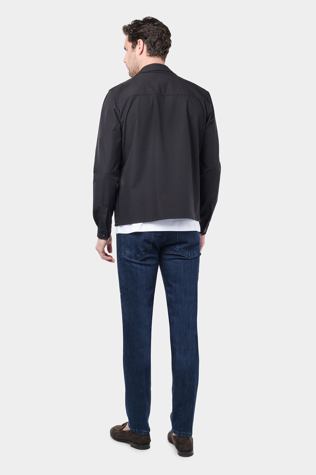 Un homme de dos vêtu d'un blouson noir en tissu Olmetex, imperméable et coupe-vent, décontracté, avec quatre poches, une fermeture à double zip et un col. Il porte un tee-shirt blanc en coton glacé et un jean couleur brut , accompagné de mocassins en daim marron.