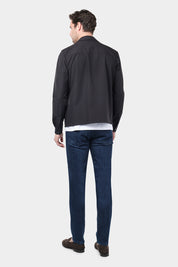 Un homme de dos vêtu d'un blouson noir en tissu Olmetex, imperméable et coupe-vent, décontracté, avec quatre poches, une fermeture à double zip et un col. Il porte un tee-shirt blanc en coton glacé et un jean couleur brut , accompagné de mocassins en daim marron.