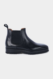 Paire de bottines Chelsea en cuir noir brillant avec des élastiques noirs sur les côtés et des languettes noires à l'arrière, posées sur un fond gris clair