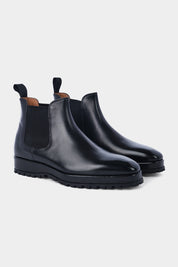 Paire de bottines Chelsea en cuir noir brillant avec des élastiques noirs sur les côtés et des languettes noires à l'arrière, posées sur un fond gris clair