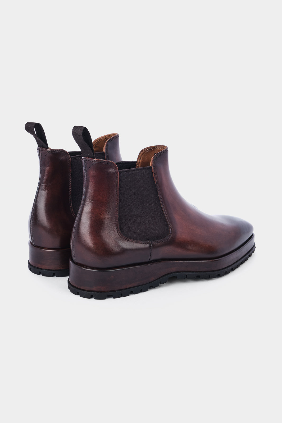 Paire de bottines Chelsea en cuir marron brillant avec des élastiques noirs sur les côtés et des languettes noires à l'arrière, posées sur un fond gris clair