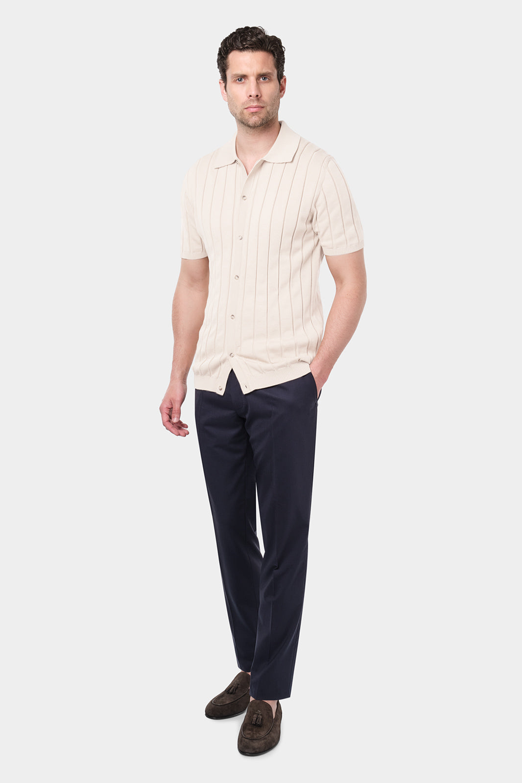 Un homme portant une chemise polo beige en coton avec des rayures verticales et un pantalon bleu marine. Il a les mains dans les poches et porte des mocassins en daim marron. Le fond est uni et de couleur claire.